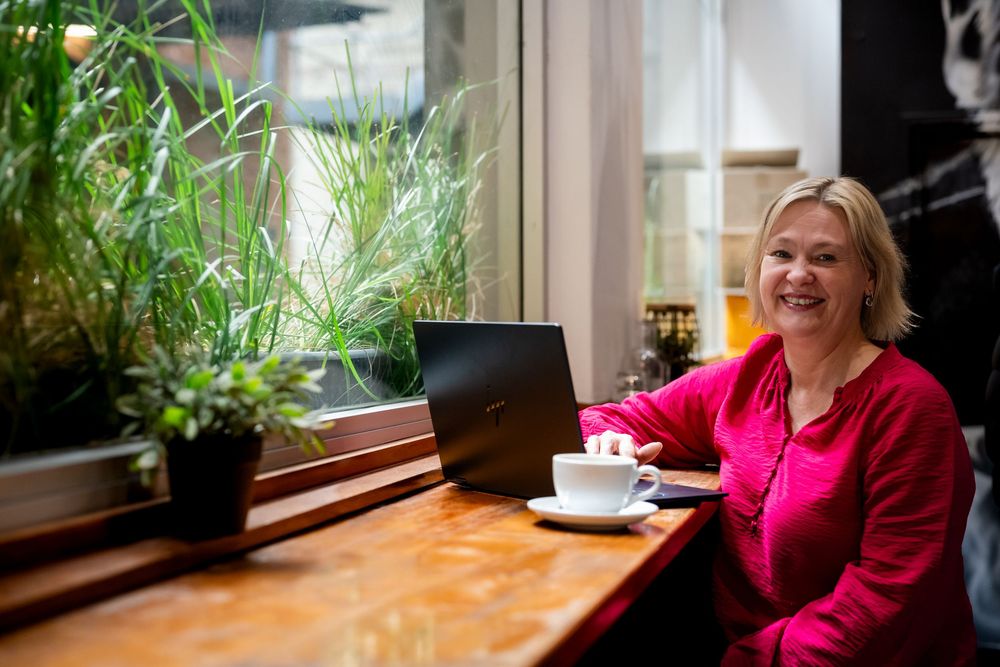 Smiling woman in red top working on laptop at cafe window table with coffee cup and green plants.