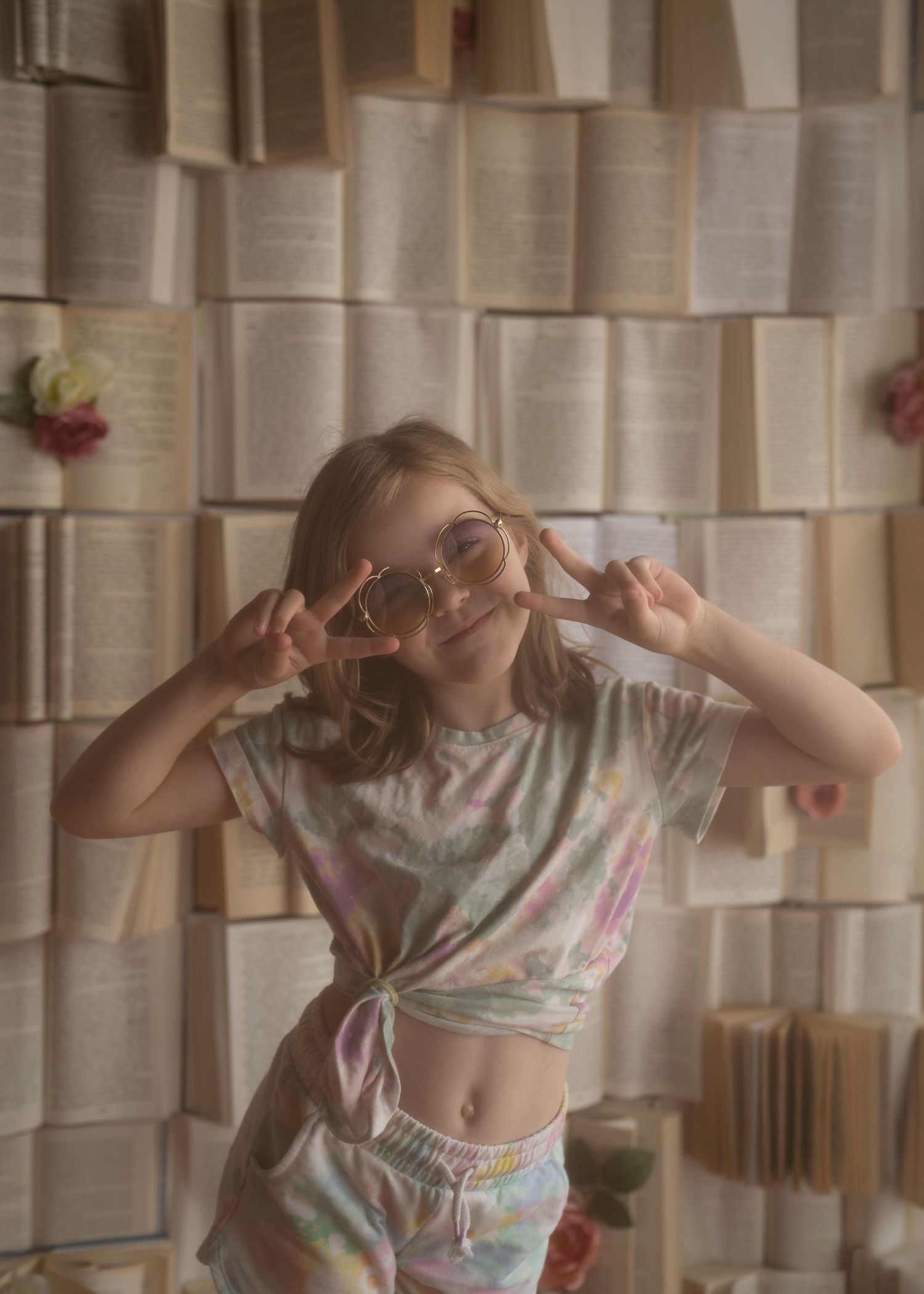 A dreamy portrait against a wall of open books showing someone in a tie-dye outfit making peace signs.