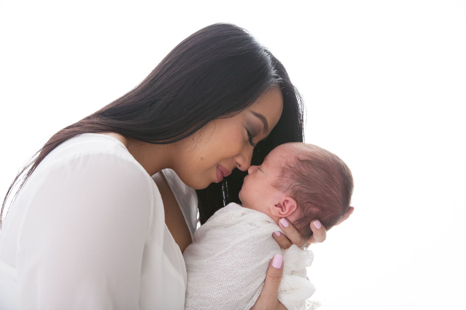 A tender moment captured as a woman in white gently holds and nuzzles a newborn baby wrapped in white fabric.
