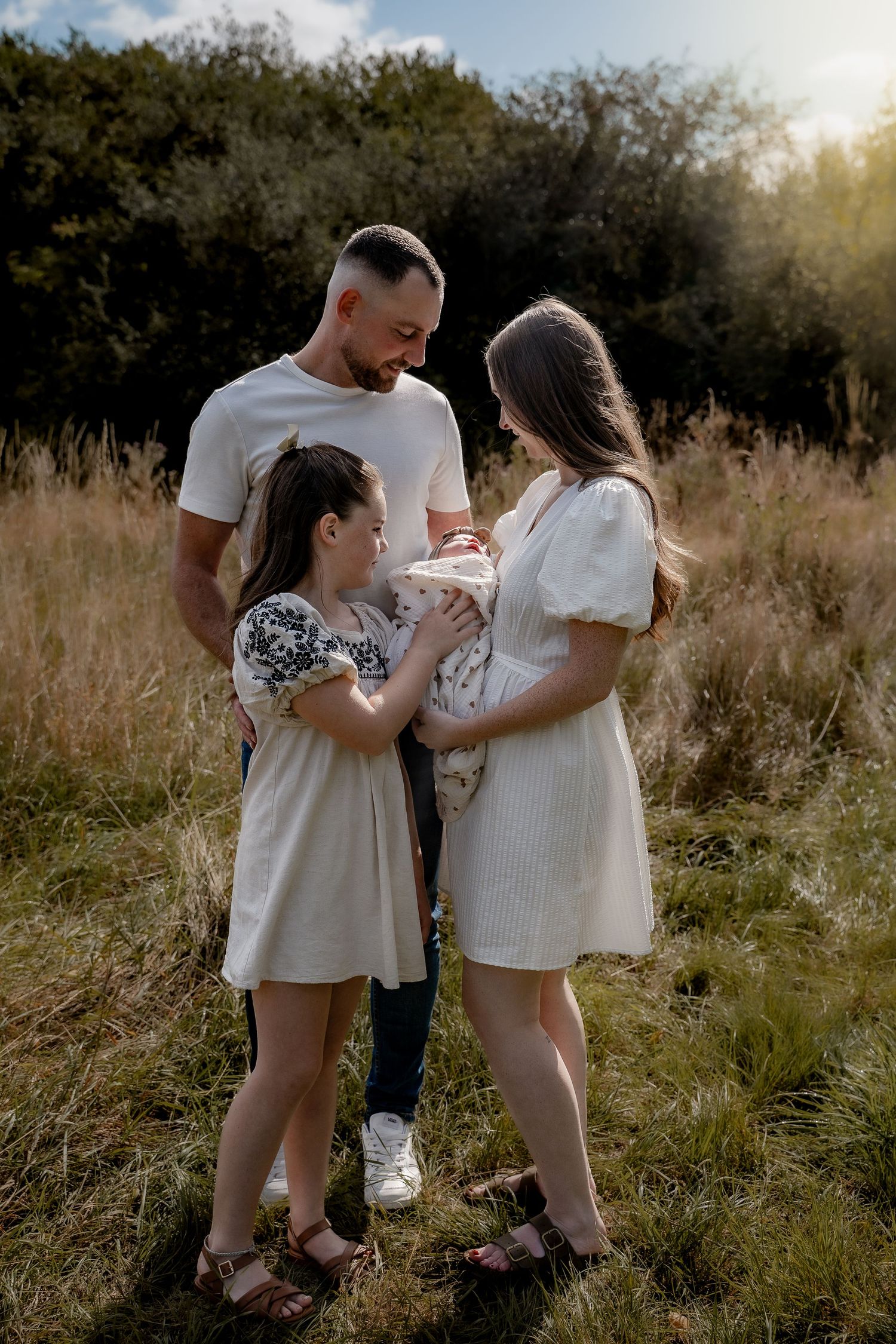 A family in white outfits shares tender moments together in a sunlit meadow during a golden hour photoshoot.