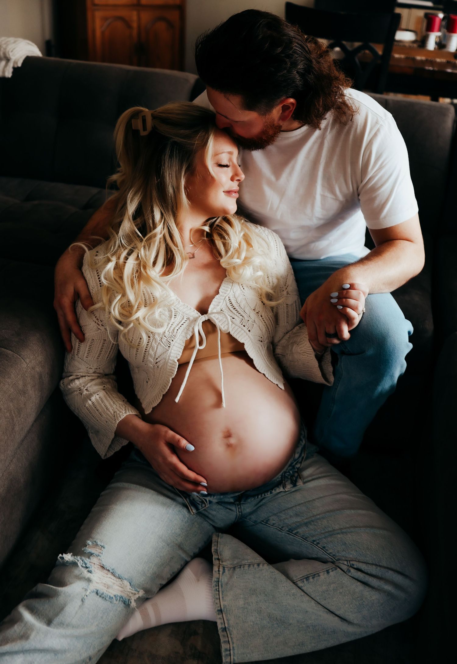 Intimate maternity photoshoot showing couple in casual attire sitting together on couch in moody lighting.