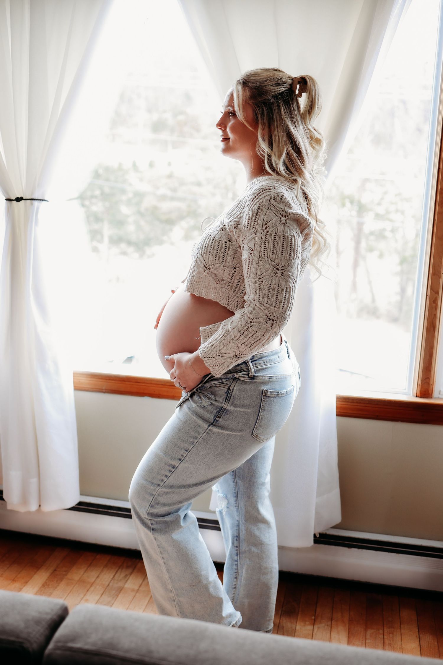Side profile views of a pregnant woman in light jeans and white sweater standing by sunlit windows.