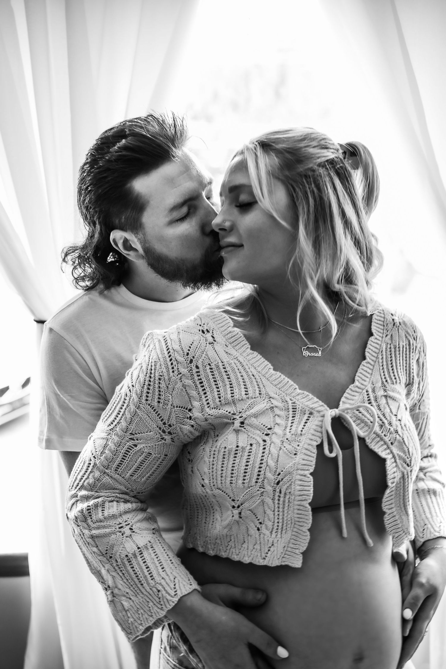 A romantic black and white maternity photo shows an intimate moment between a couple in front of sheer curtains.