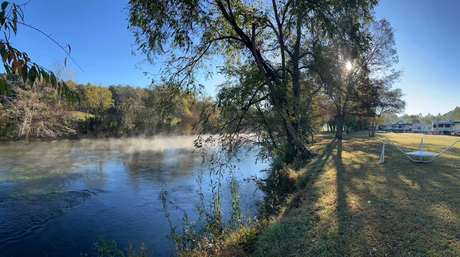 Morning mist rises from a calm river beside sunlit trees in a peaceful autumn landscape.