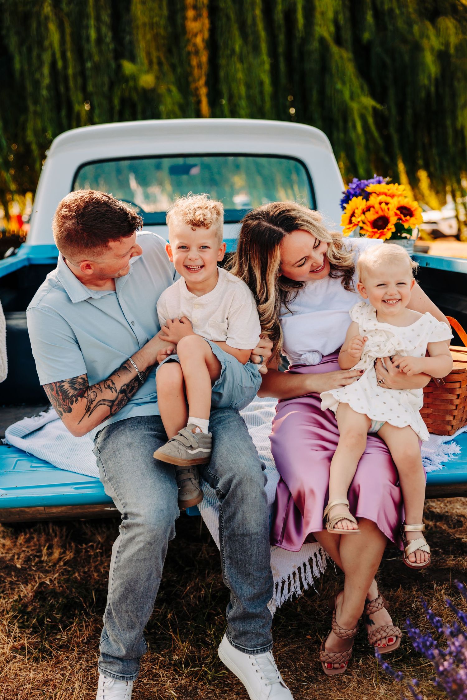 A family sits together on the back of a vintage blue pickup truck surrounded by sunflowers on a sunny afternoon.