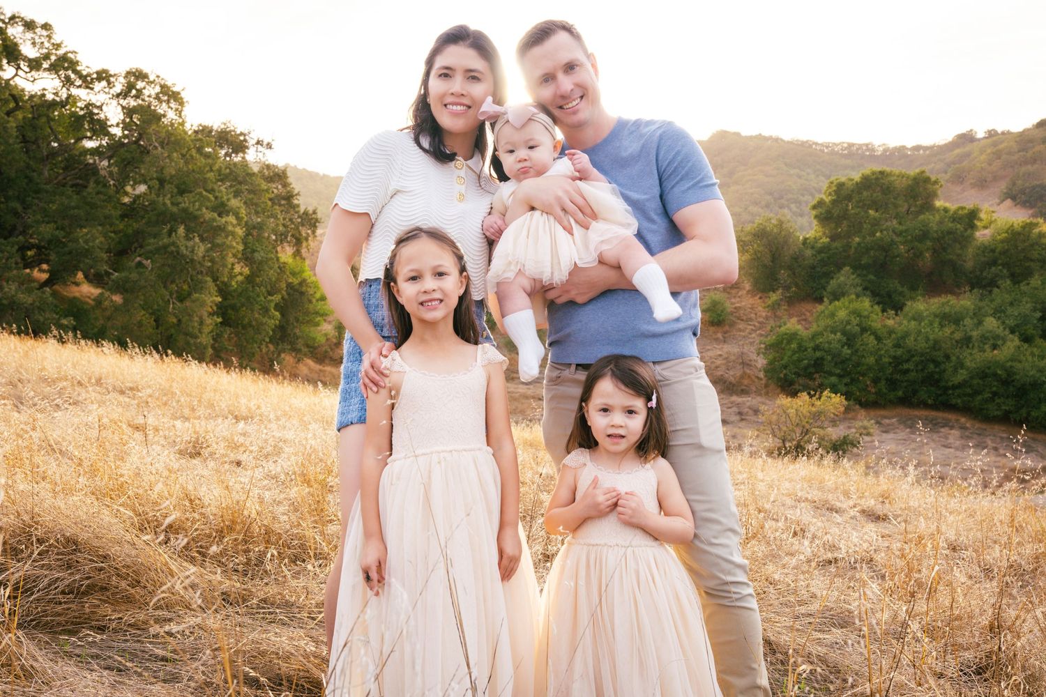A family in light neutral outfits poses together in a golden field during sunset for outdoor portraits.