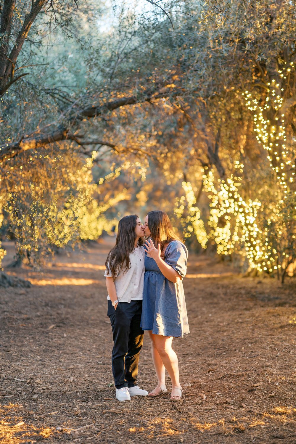 Two people share an intimate moment on a path surrounded by glowing trees at dusk.
