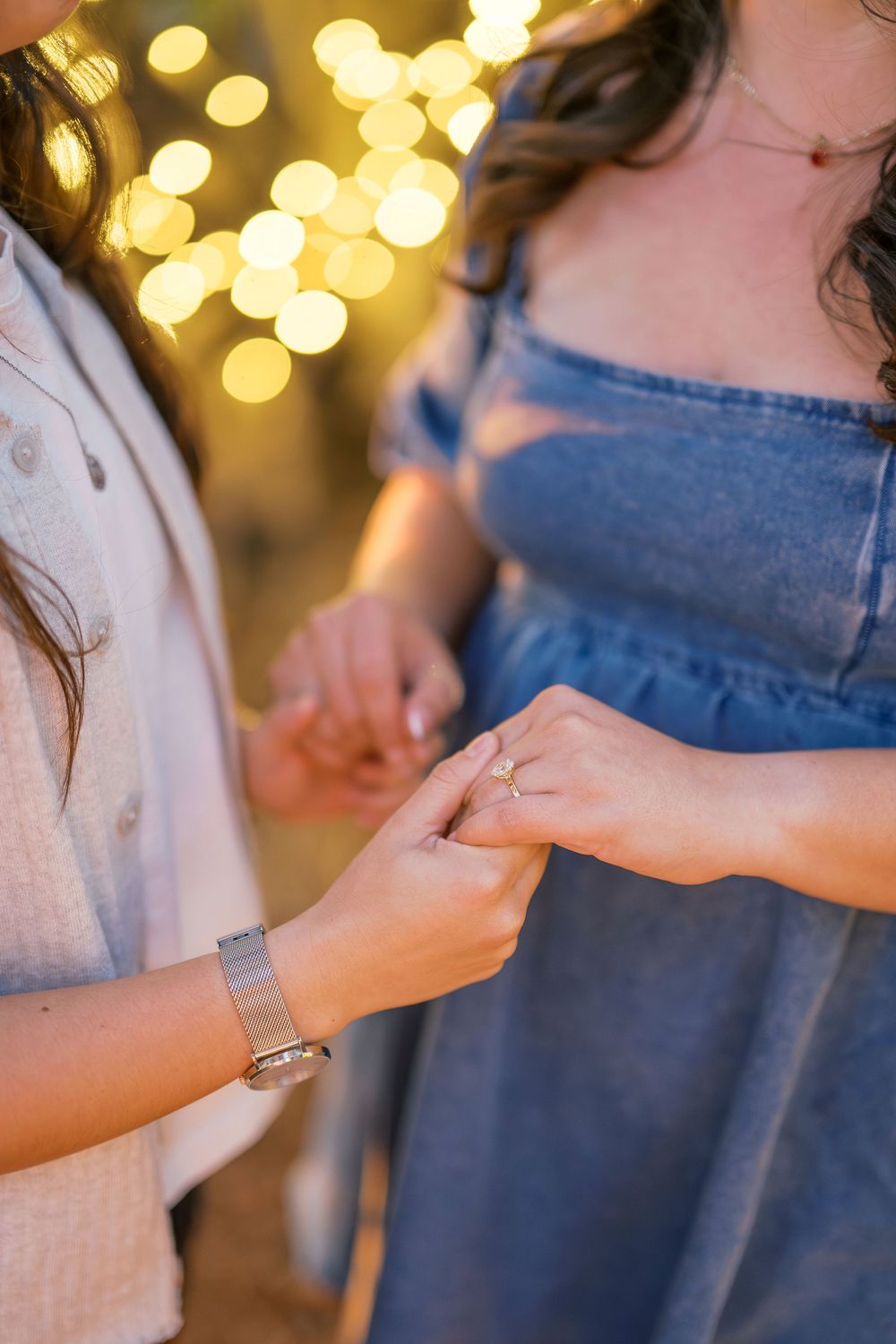 Two people hold hands with romantic bokeh lights in the background as one wears denim and the other white.