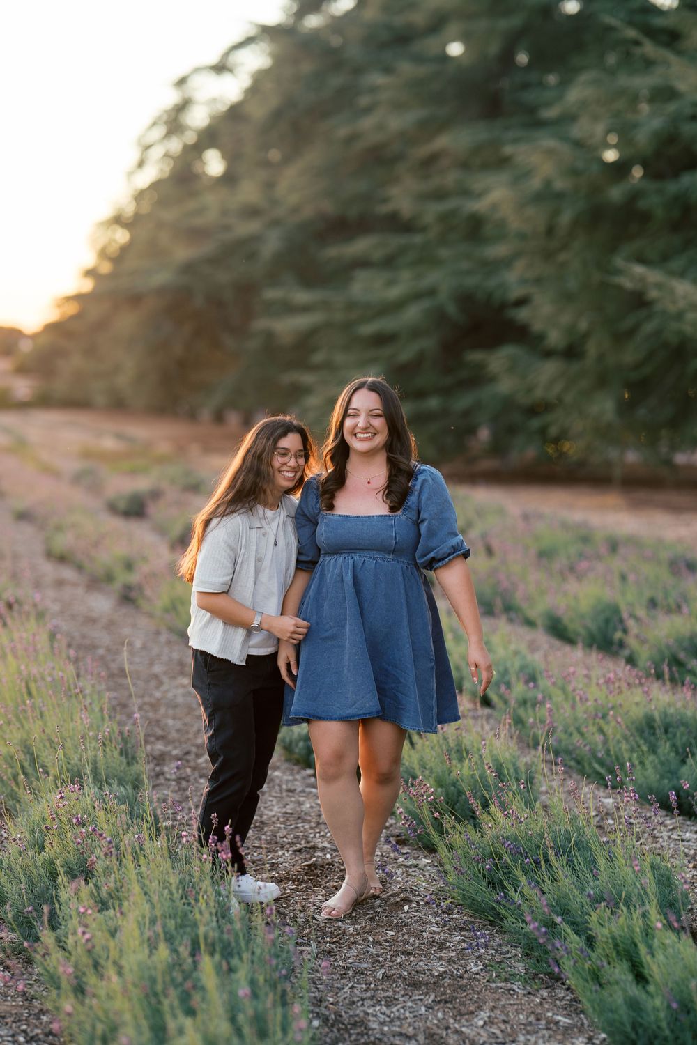 Two friends walk together and laugh on a dirt path through blooming purple lavender fields at sunset.