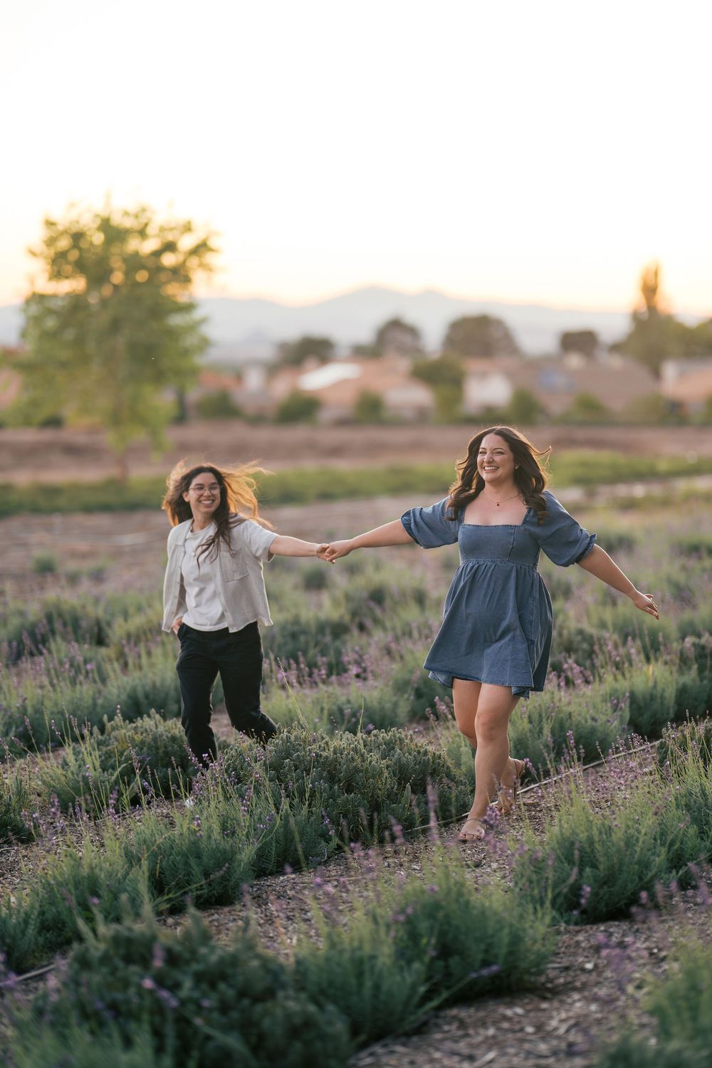 Two people dance playfully through a lavender field at sunset with mountains in the background.