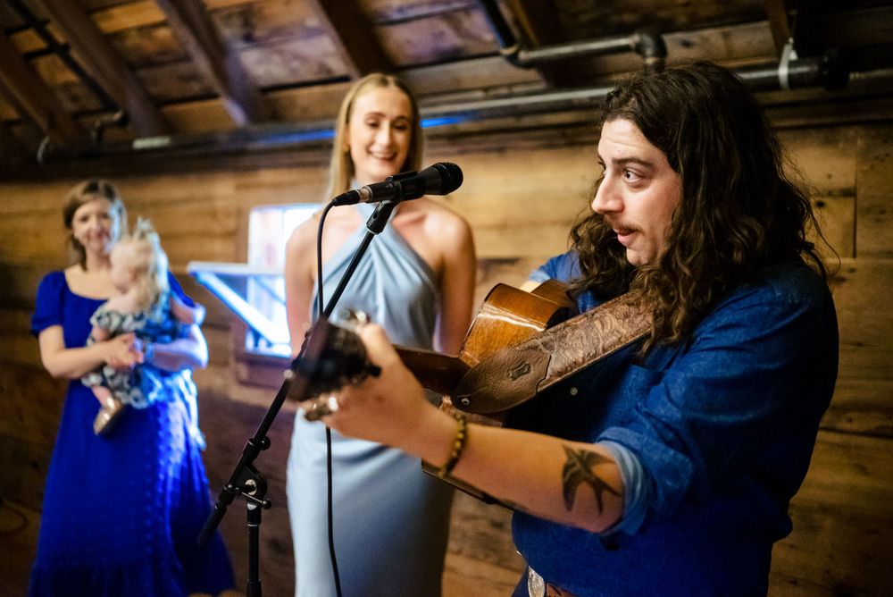 Tattooed guitarist performing at a rustic barn wedding with bridesmaids watching nearby.