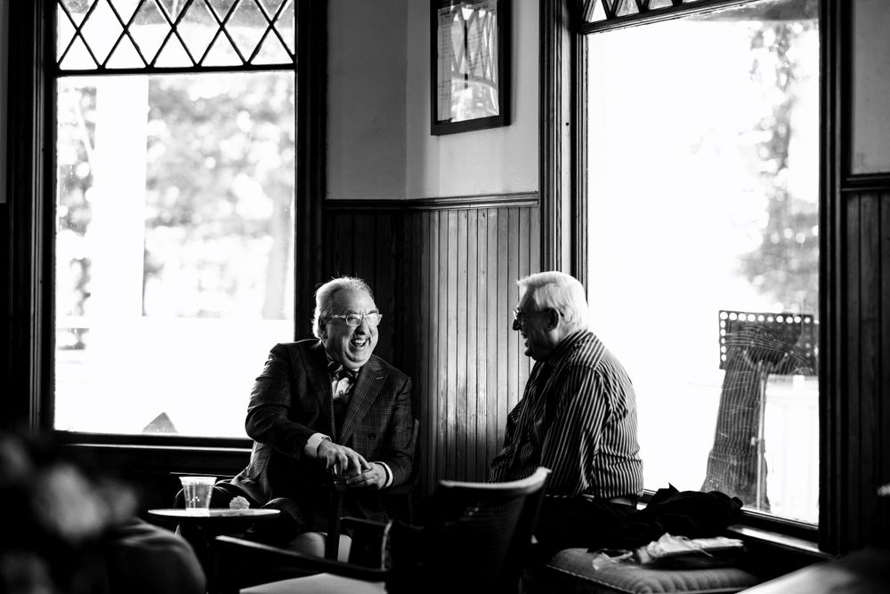 Two elderly men share a candid conversation at a warmly lit pub table in black and white photography.