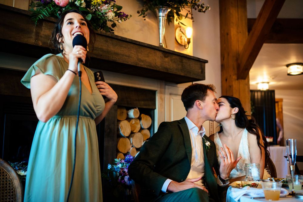 A bridesmaid delivers a heartfelt speech while the bride and groom share a kiss at the reception.
