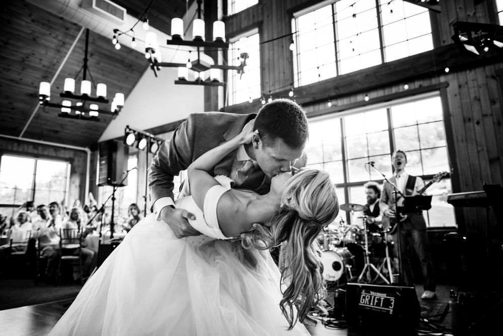 Groom dips bride dramatically during their first dance at a rustic wedding venue in black and white.