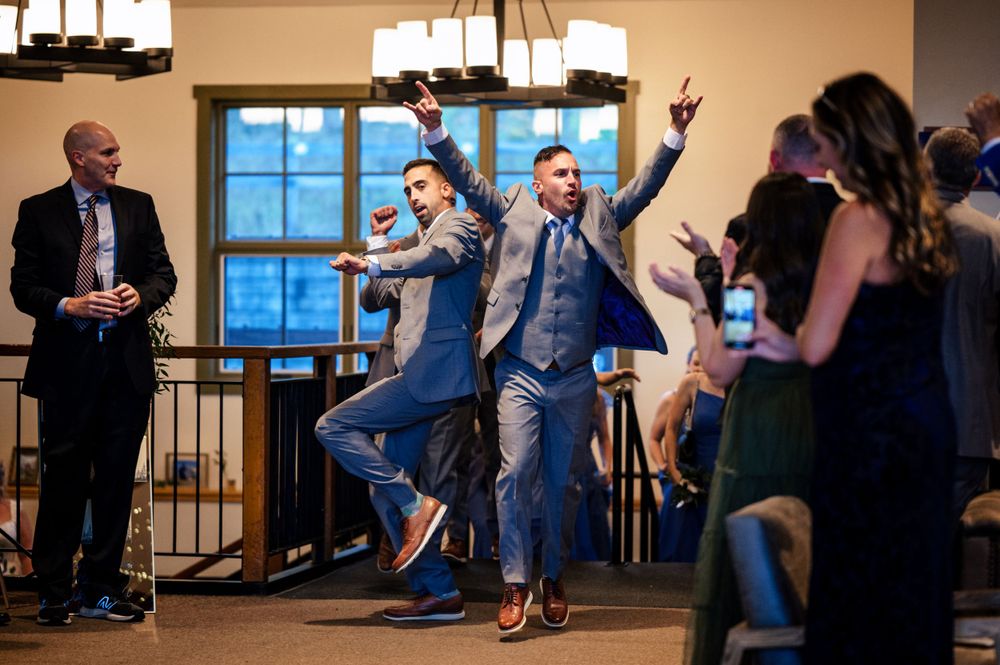 Two men in blue suits dance energetically at indoor wedding reception venue.