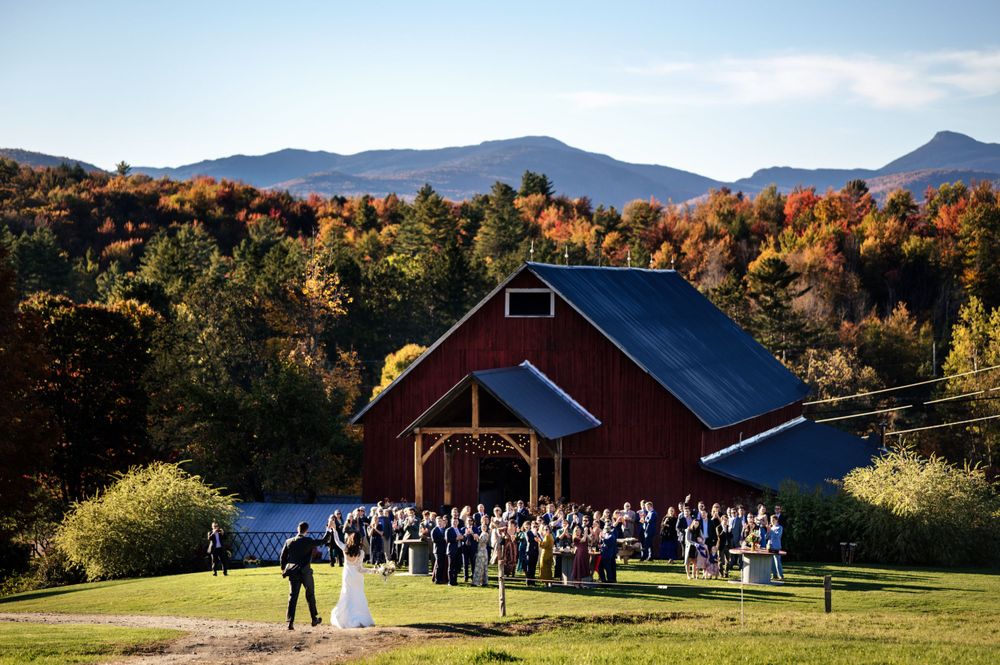 Outdoor wedding ceremony with guests gathered before a red barn, framed by vibrant fall foliage and mountain views.