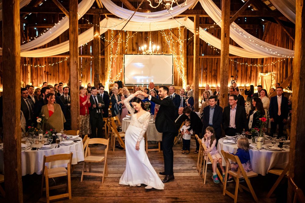 Bride and groom share first dance in warmly lit rustic barn decorated with string lights and draped fabric.