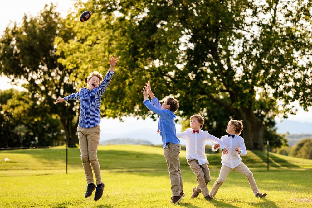Four boys in dress shirts joyfully jump and play catch with a hat on a sunny green lawn.