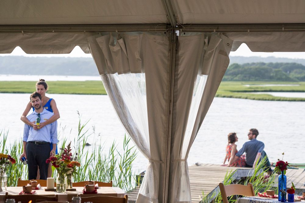 Wedding guests mingle under an elegant white tent overlooking a serene waterfront marsh landscape.