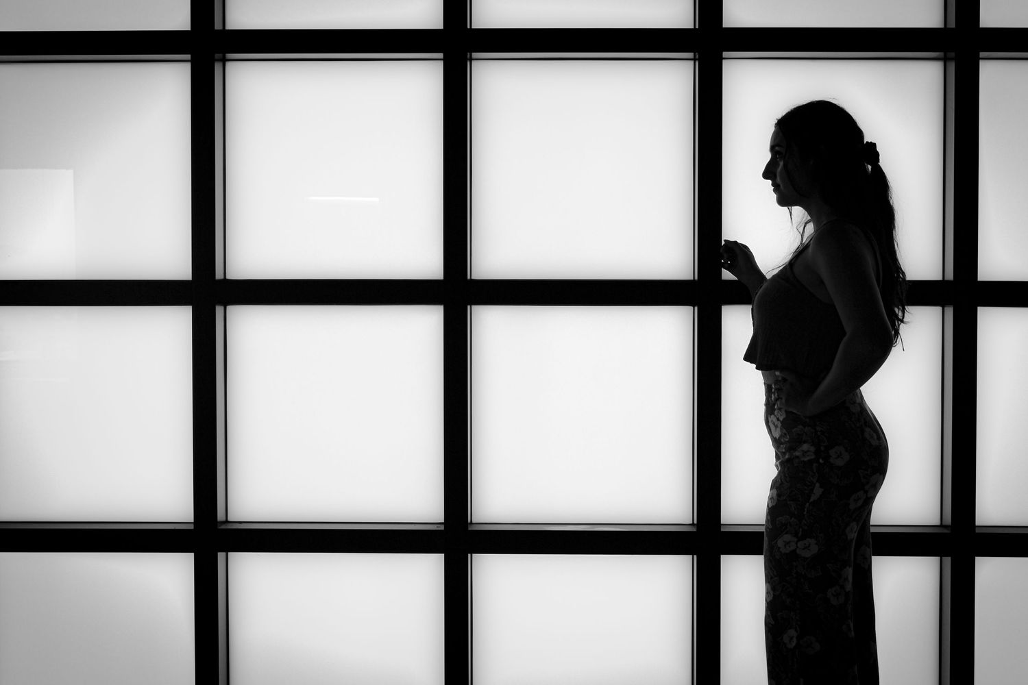 Silhouette of a woman standing indoors against a large grid-patterned backlit window, side profile, hair in ponytail, wearing floral pants.