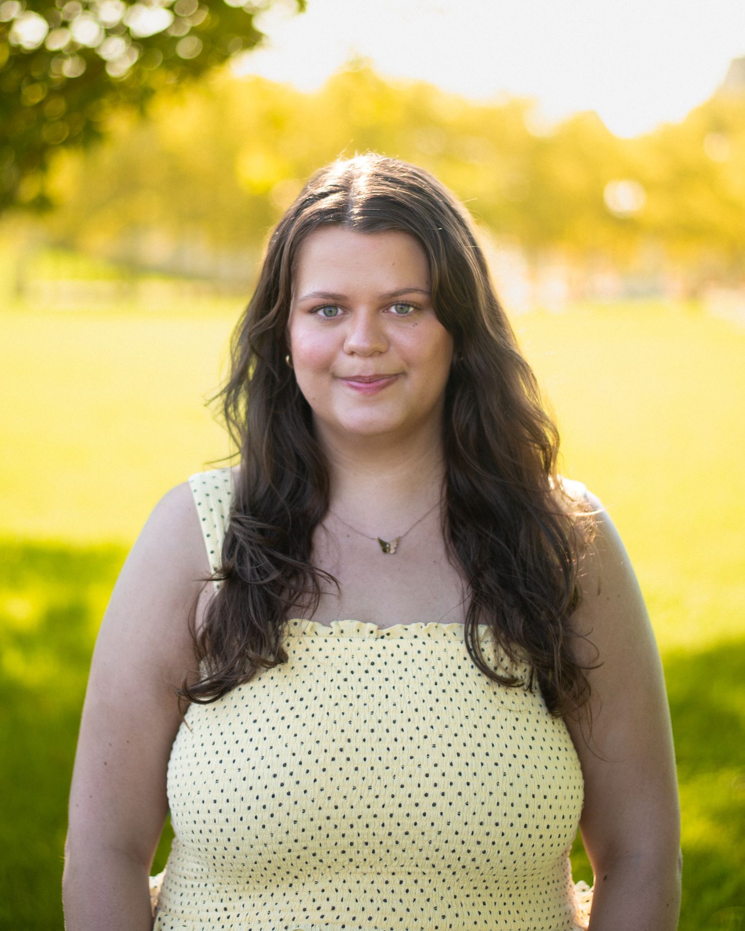 Young woman with long dark hair wearing a yellow polka dot sundress, smiling outdoors in a sunny green park.
