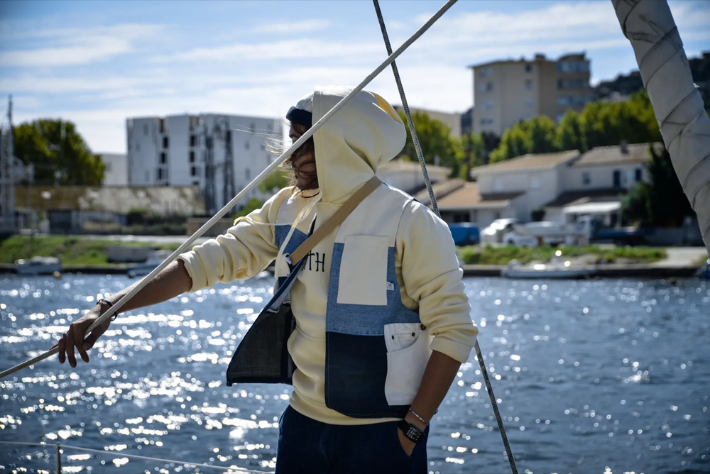 Homme sur un voilier en rivière urbaine, tendant une corde, habillé en beige et bleu, avec un décor d’immeubles et de verdure.