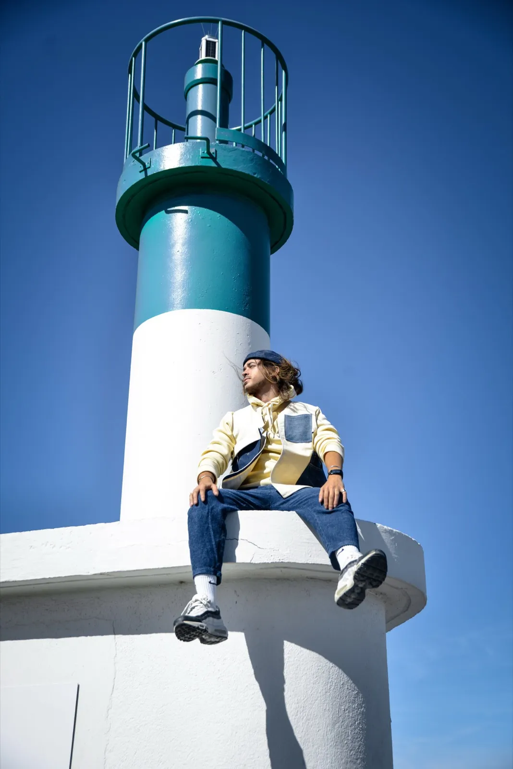 Personne assise au sommet d’un phare blanc et vert, dominant un ciel bleu vif, posture détendue et contemplative.