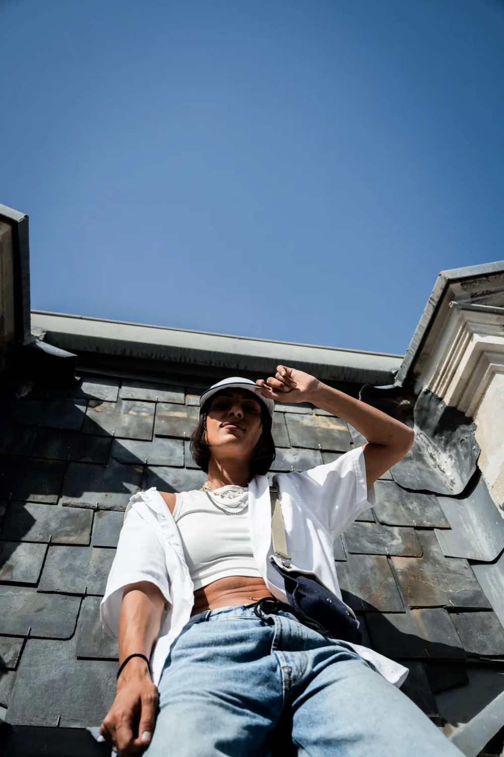 Femme assise sur un toit en ardoise, main sur le front, vêtue d’un crop top blanc et jean.