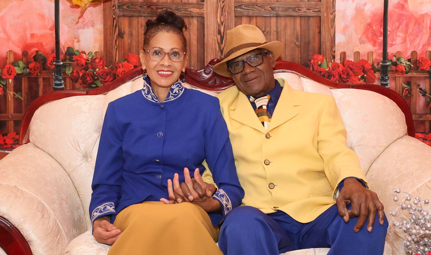 A couple poses together in coordinating blue and yellow outfits against a pink floral backdrop.