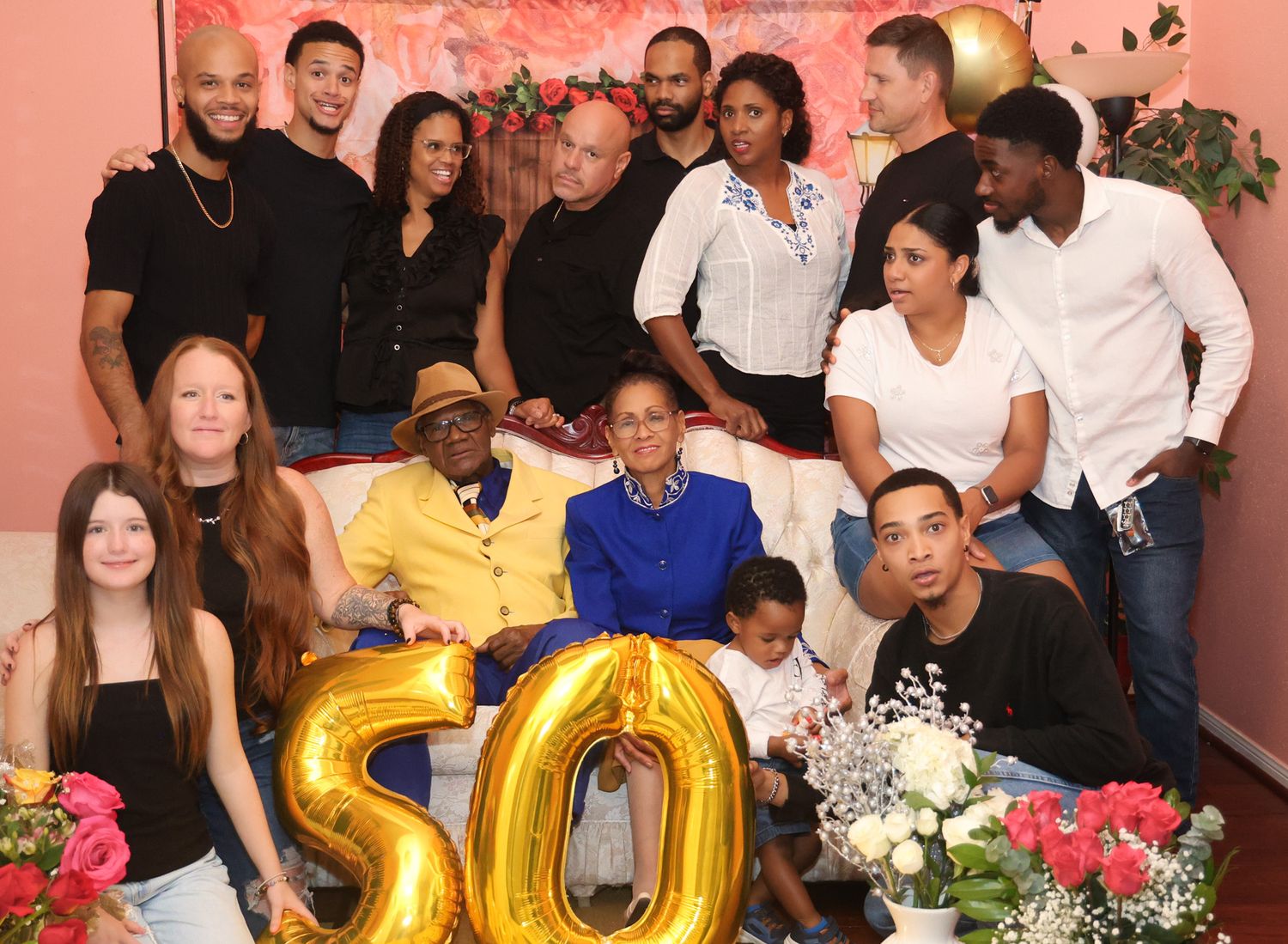 50th birthday celebration group photo with gold balloons and pink floral backdrop.