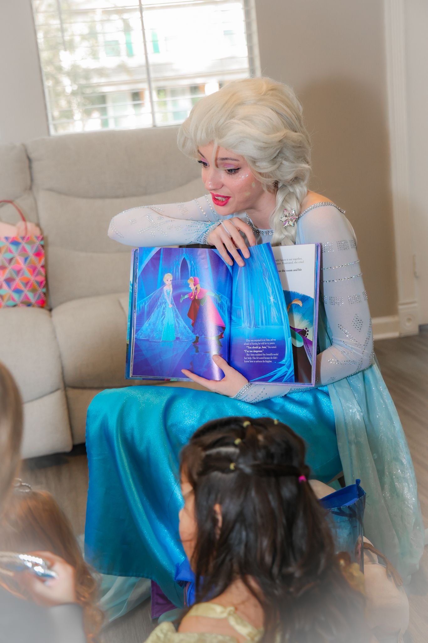 A performer in a sparkly blue dress reads from a storybook while sitting on a couch.
