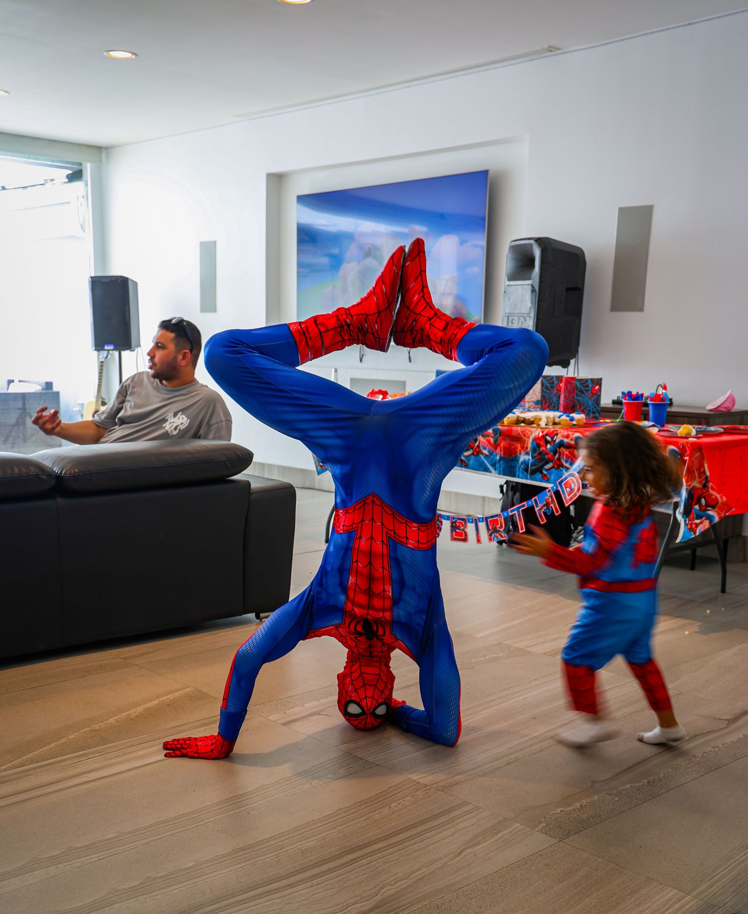 Spider-Man performer demonstrating athletic moves in a modern indoor space with dark couches.