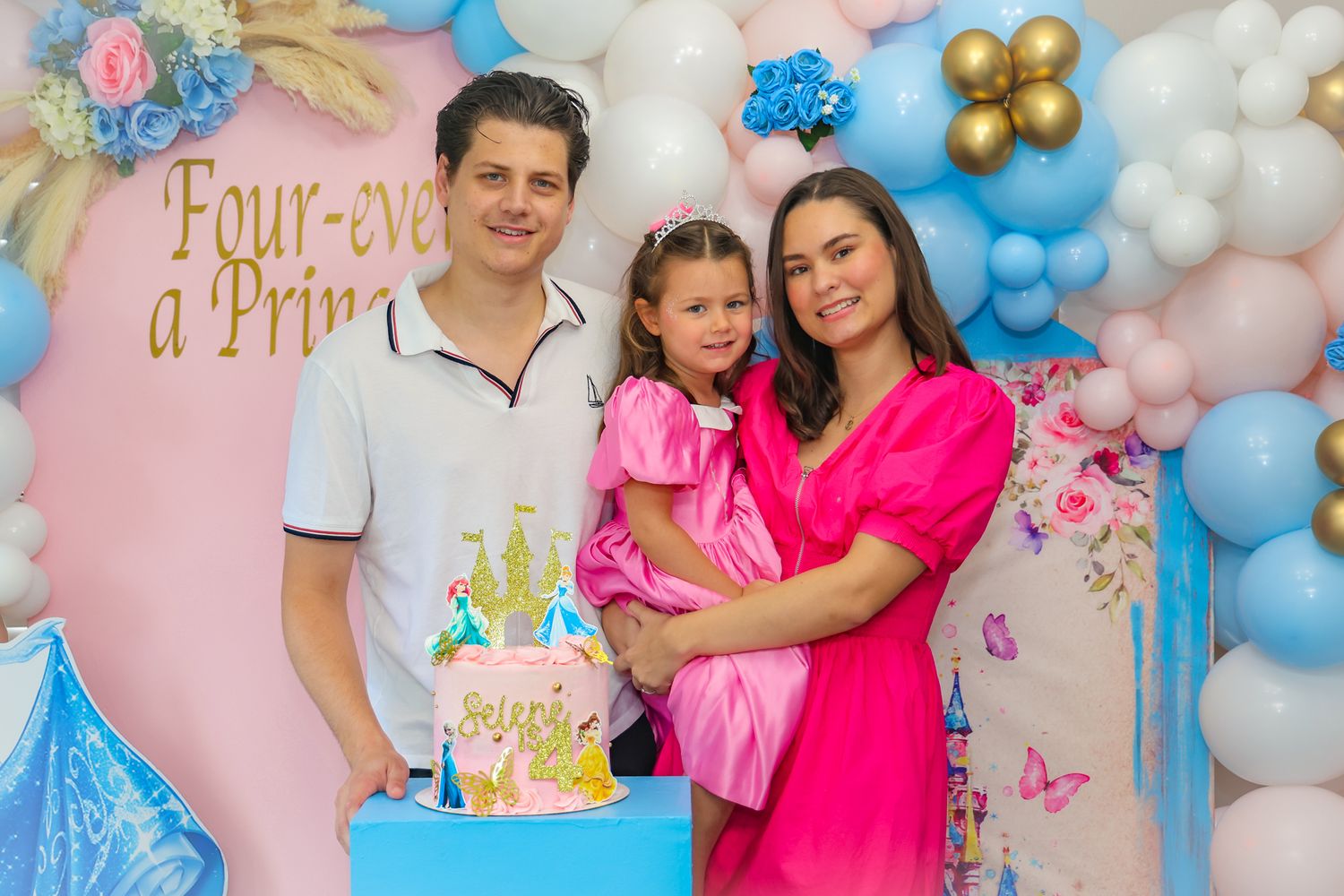 A birthday celebration with a blue and pink balloon arch backdrop and a castle-themed cake on a blue stand.