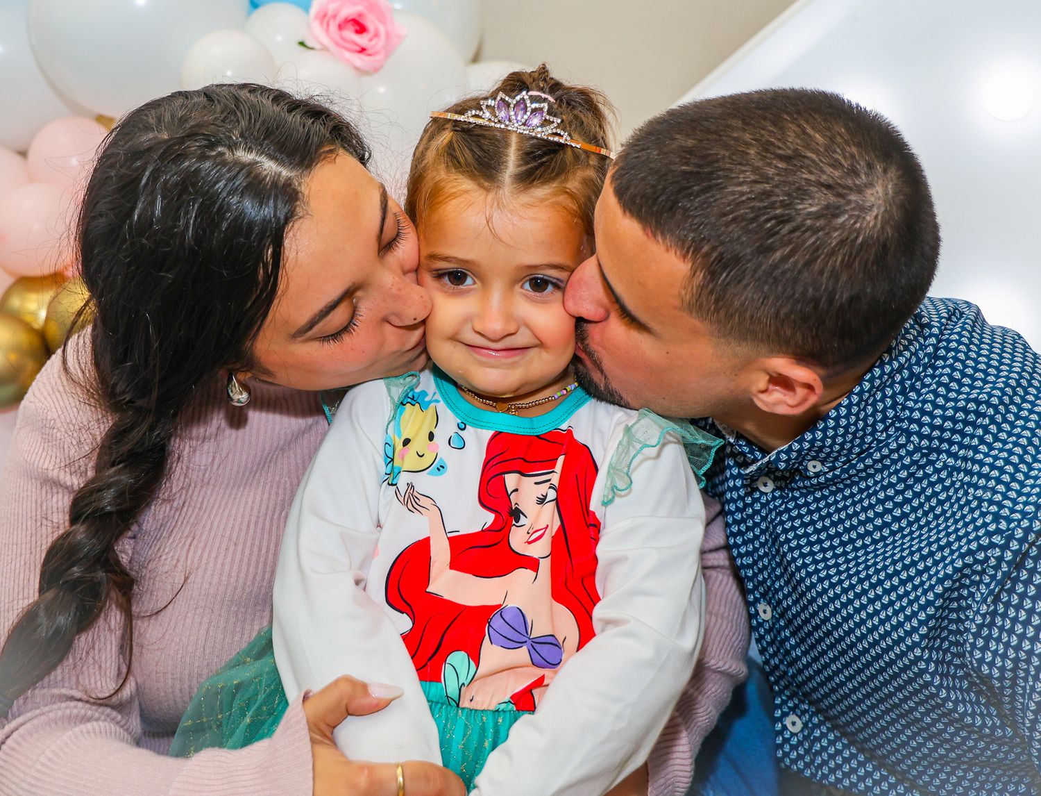 A child wearing a Little Mermaid shirt and tiara receives loving kisses from both sides against a pastel balloon backdrop.