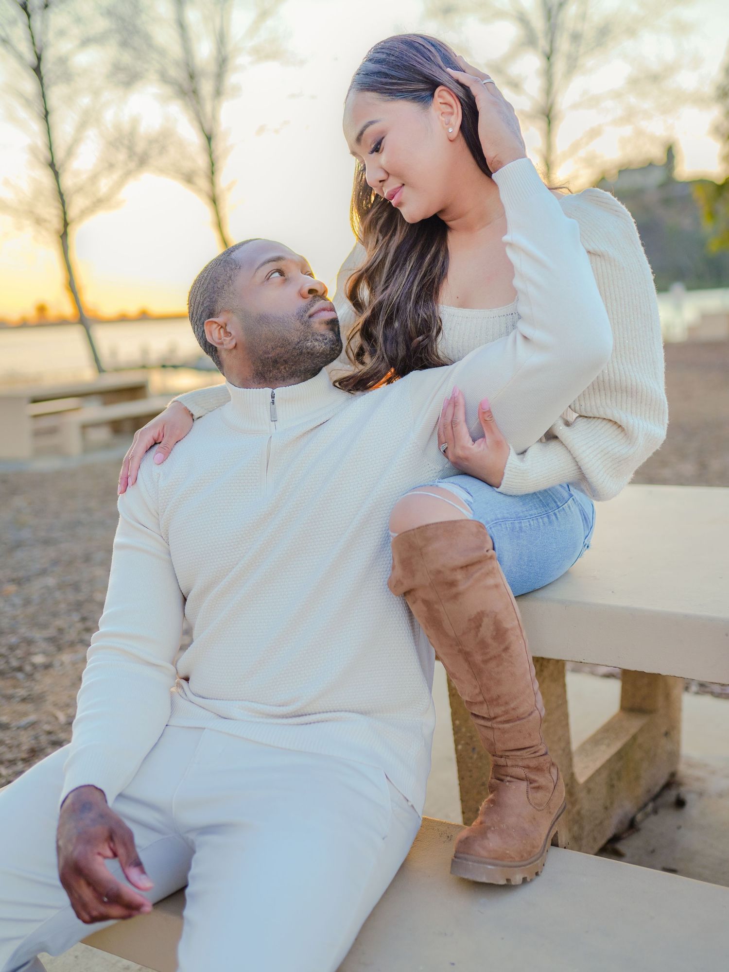 A romantic couple in coordinating white outfits share a tender moment on a wooden bench at sunset.