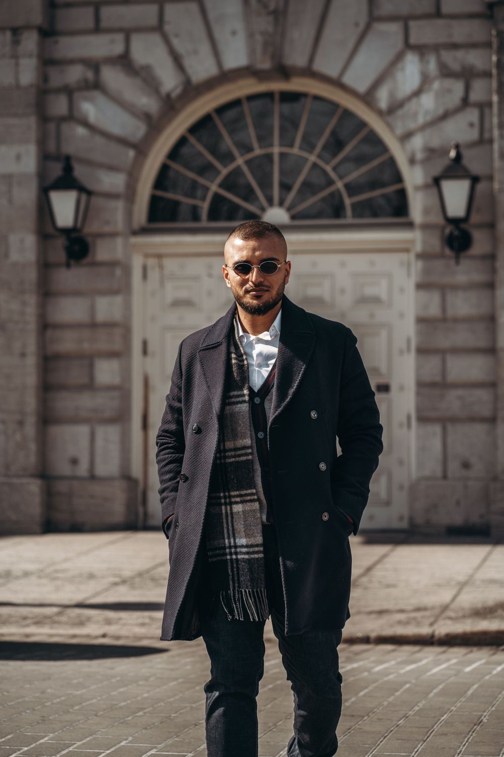 Stylishly dressed person in a dark coat and plaid scarf poses in front of an elegant arched doorway with wall lamps. Mtlshutter photography