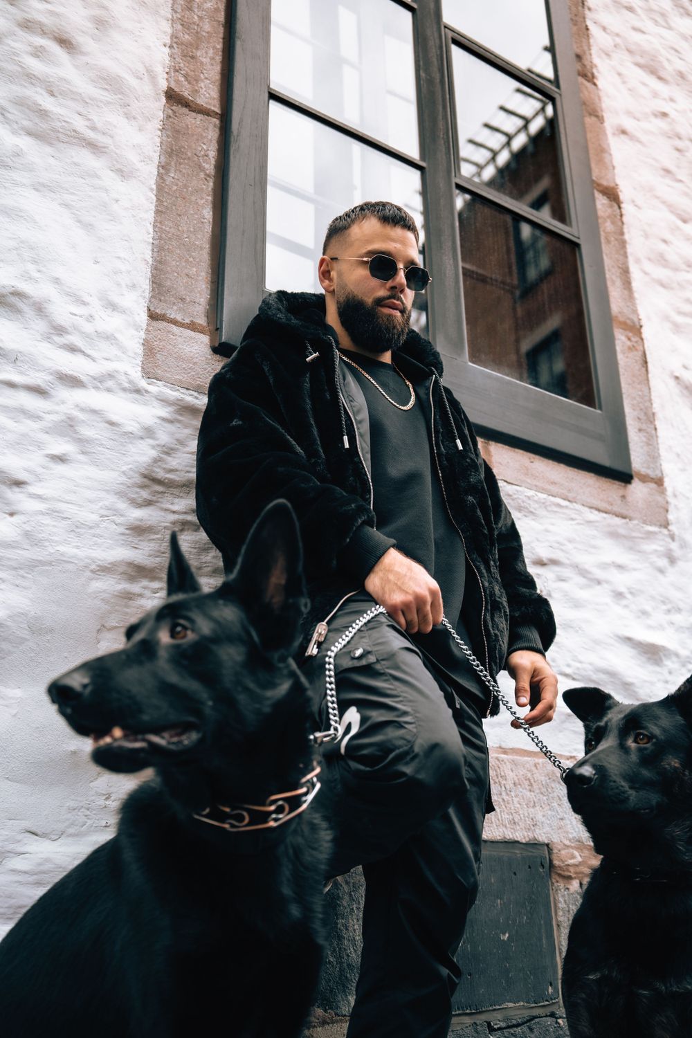 Person in black winter wear sits with two black dogs against a white building wall. Mtlshutter photography