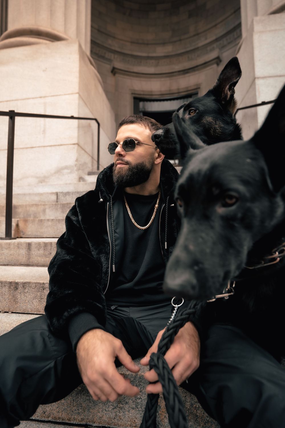 Person in black jacket sits on stone steps with two black dogs beside classical architecture. Mtlshutter photography