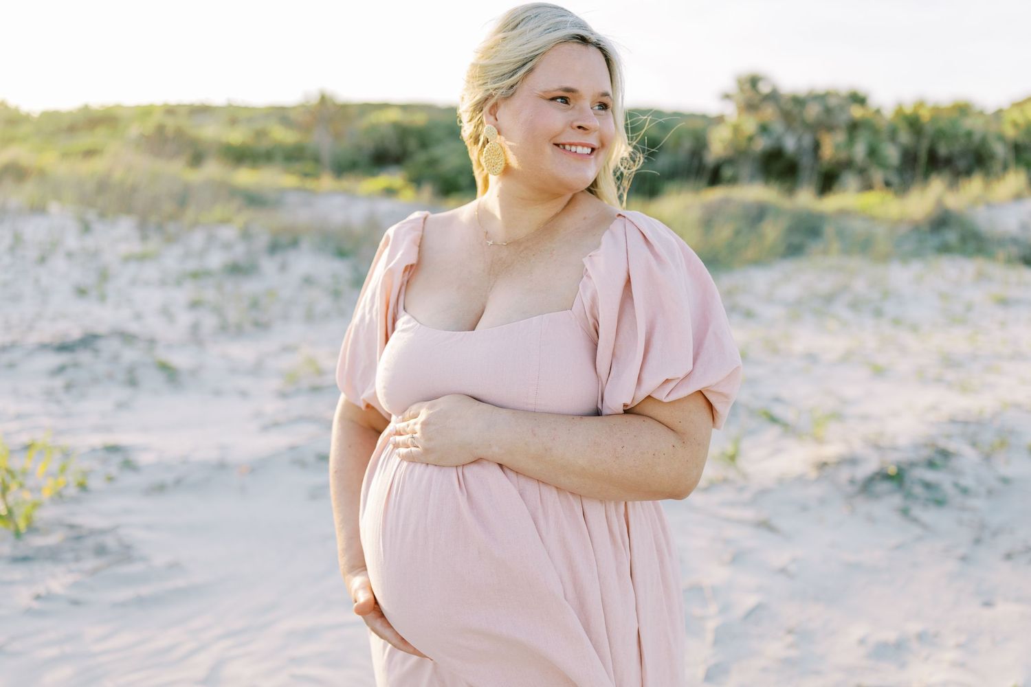 Expectant mother in black dress stands in garden with vibrant pink flowers blooming behind her.