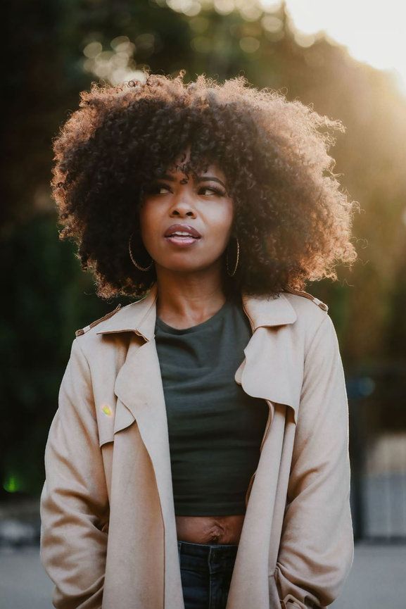 Natural curly hair catches golden sunlight while wearing a beige trench coat and crop top.