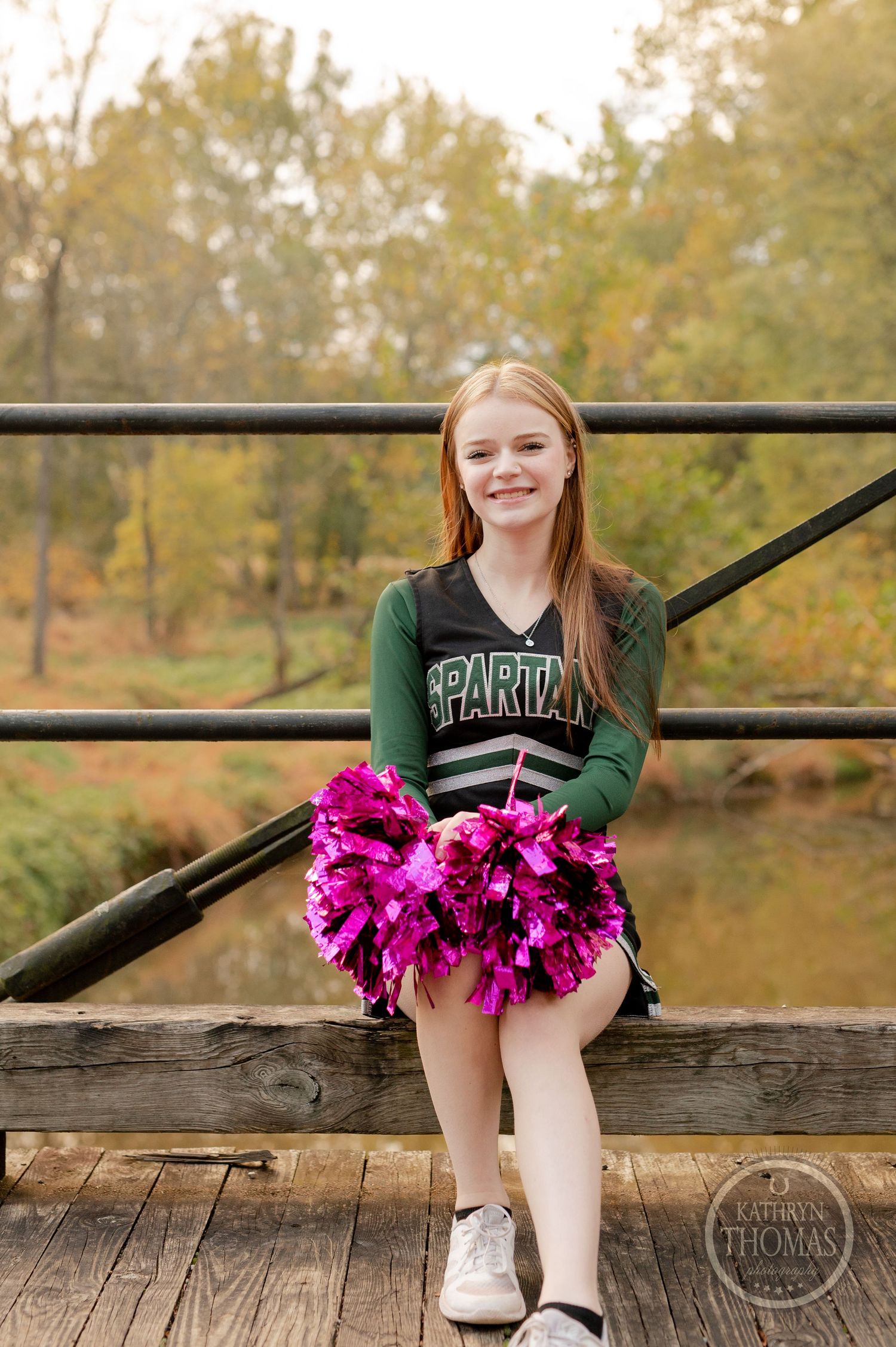 Cheerleader sits on bridge railing holding bright pink pom poms with autumn scenery in background.