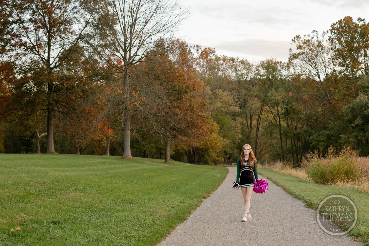 Cheerleader walks along autumn path carrying pink pom poms.