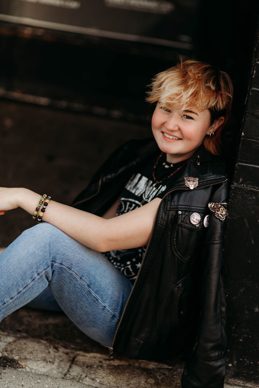 Person in leather jacket and jeans sits casually against dark wall wearing punk-style accessories.