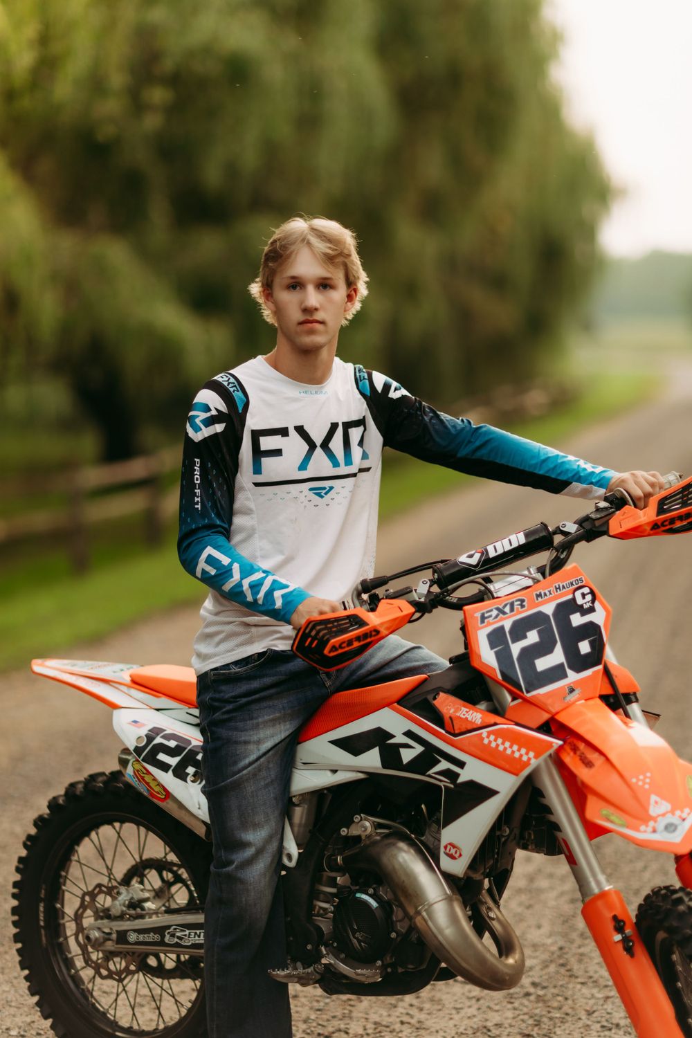 Motocross rider poses with orange dirt bike on rural road wearing FXR racing gear.