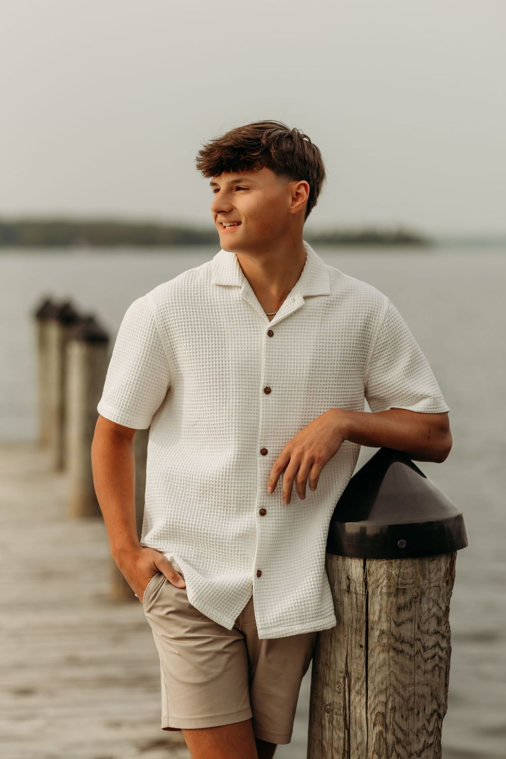 Senior in white textured shirt posing against weathered dock post at waterfront.