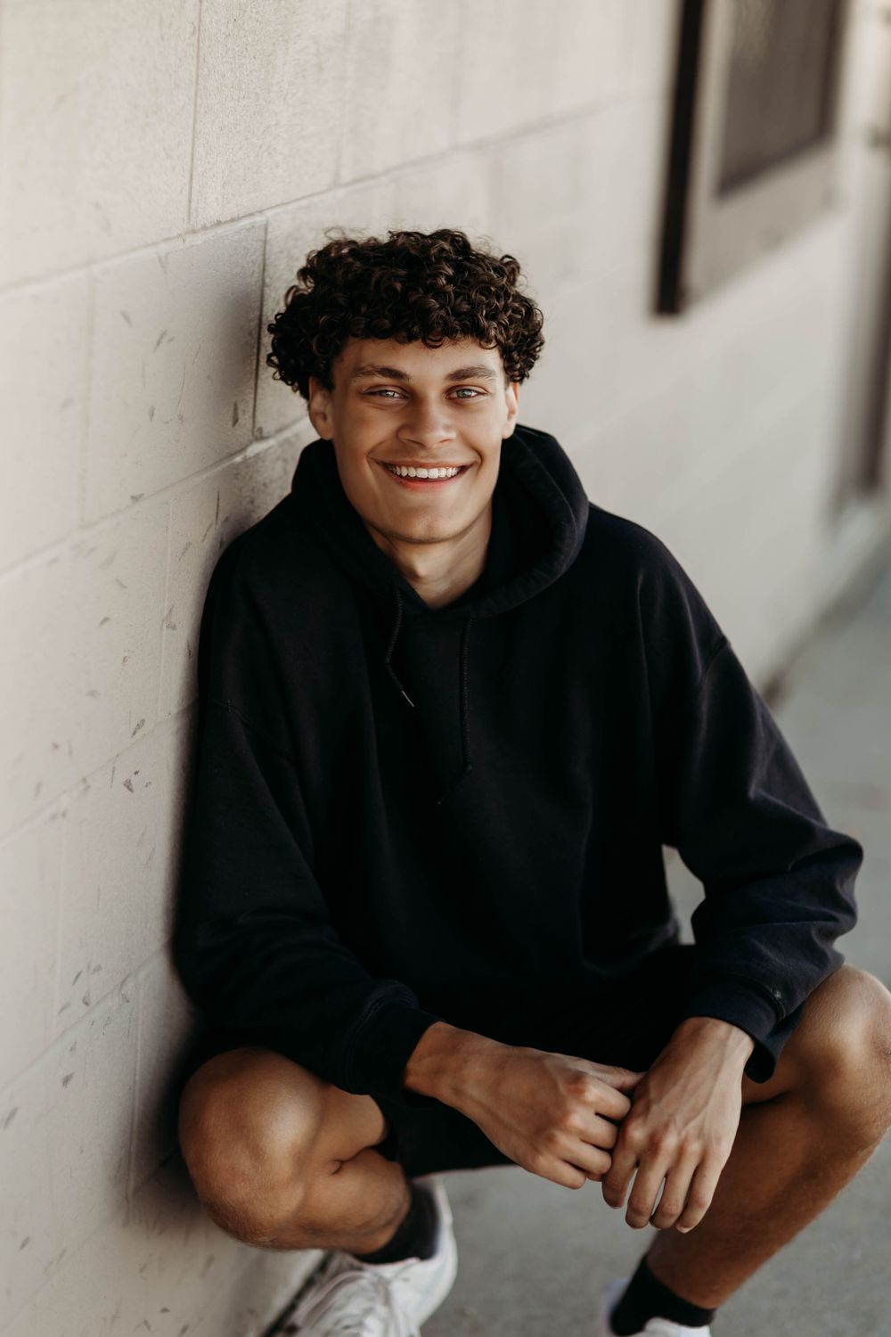 Portrait of someone in black hoodie sitting against white wall with curly hair.