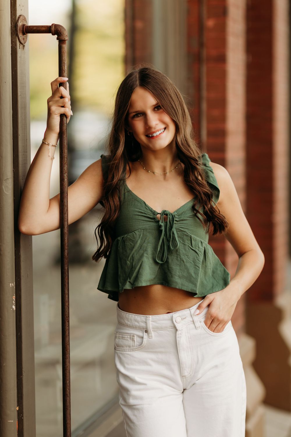 Woman in green crop top and white pants poses casually against brick wall in natural lighting.