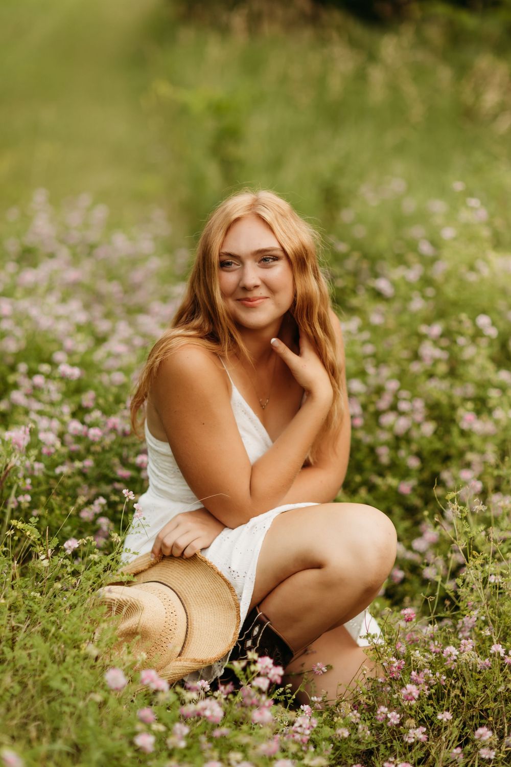 A Senior in a white dress sits in a wildflower meadow on a sunny summer day with a straw hat nearby.
