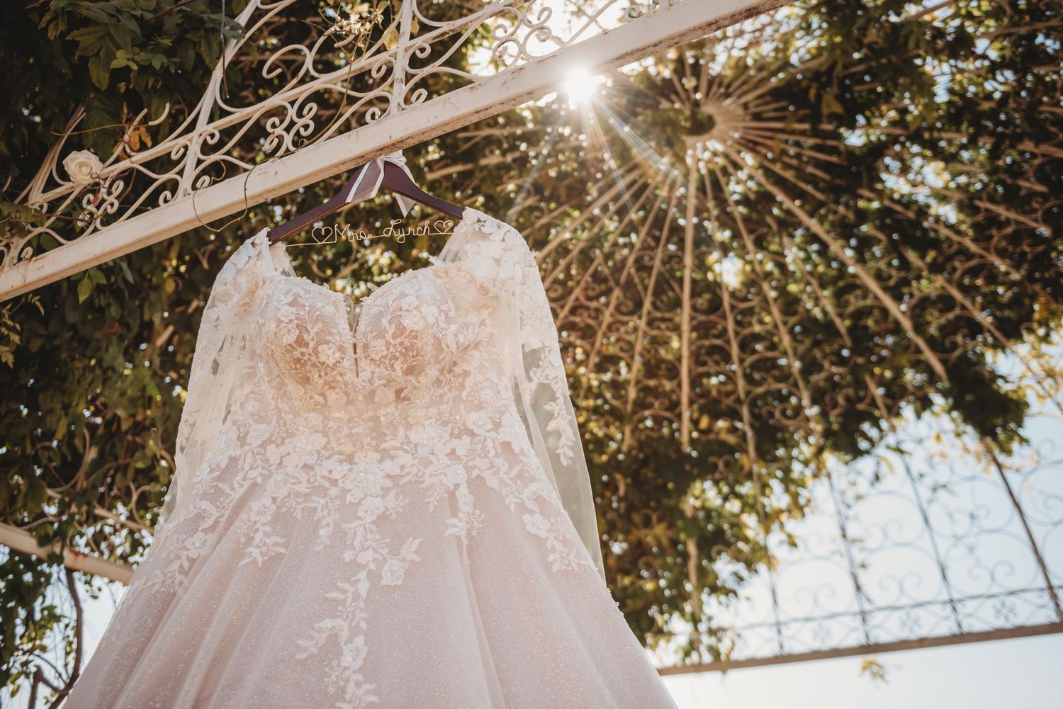 A white lace wedding dress hangs from an ornate white garden arch with sunlight streaming through tree branches above.
