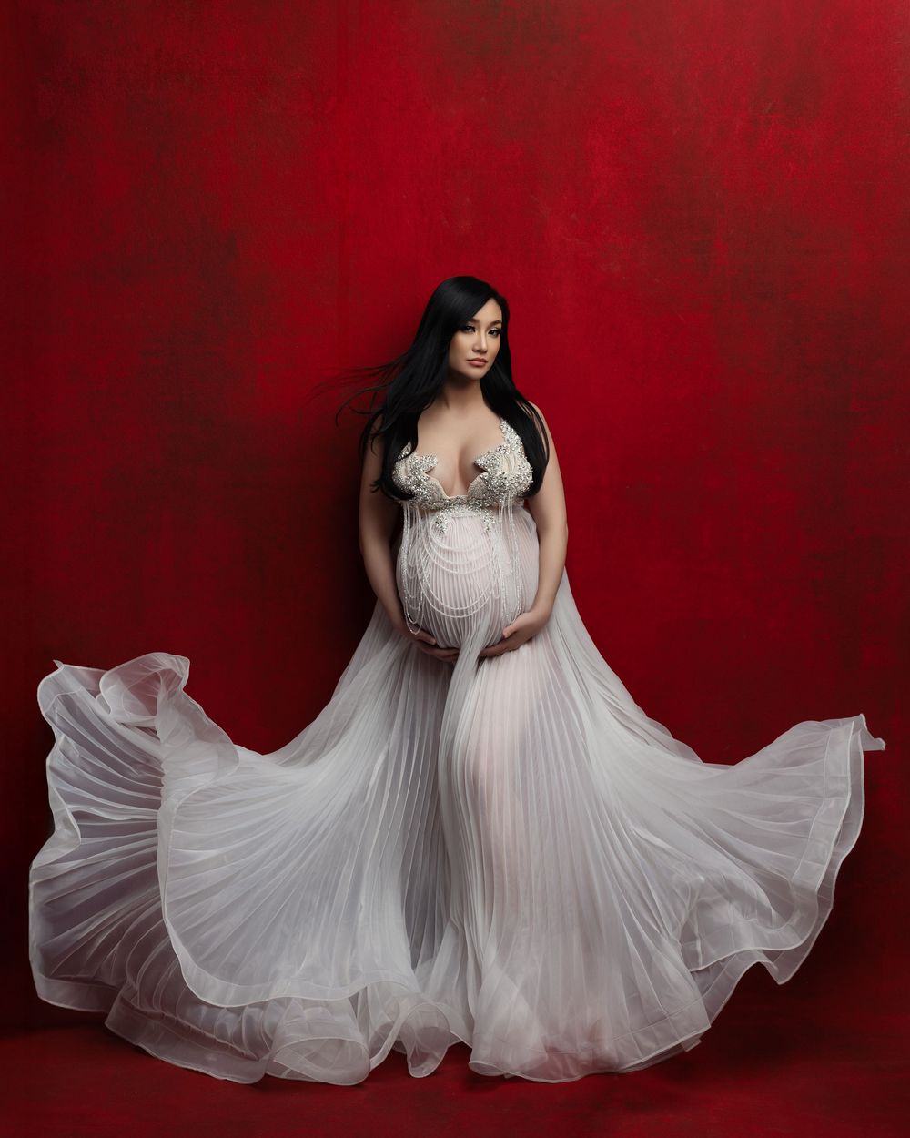 Elegant long white pleated maternity gown against deep red backdrop in artistic portrait.